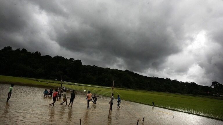 Young people play volleyball in a flooded field in India during the summer monsoon. The sky is full of dark clouds.